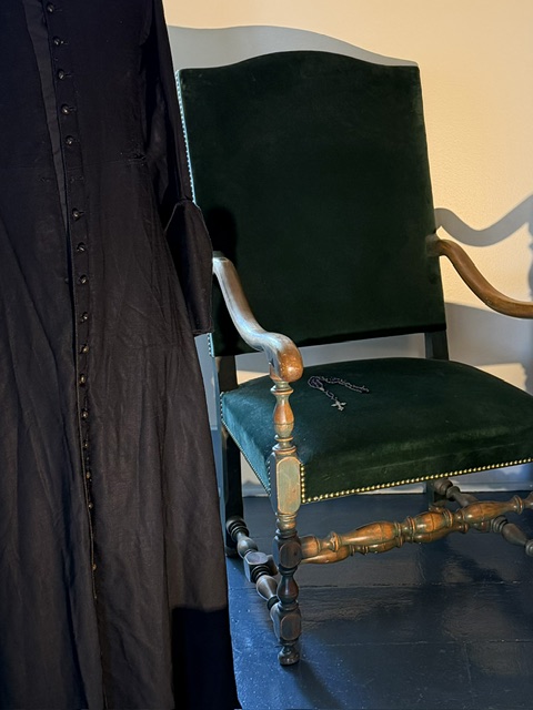 Fauteuil ancien recouvert de velours vert, près de vêtements et d’objets présentés au musée.
